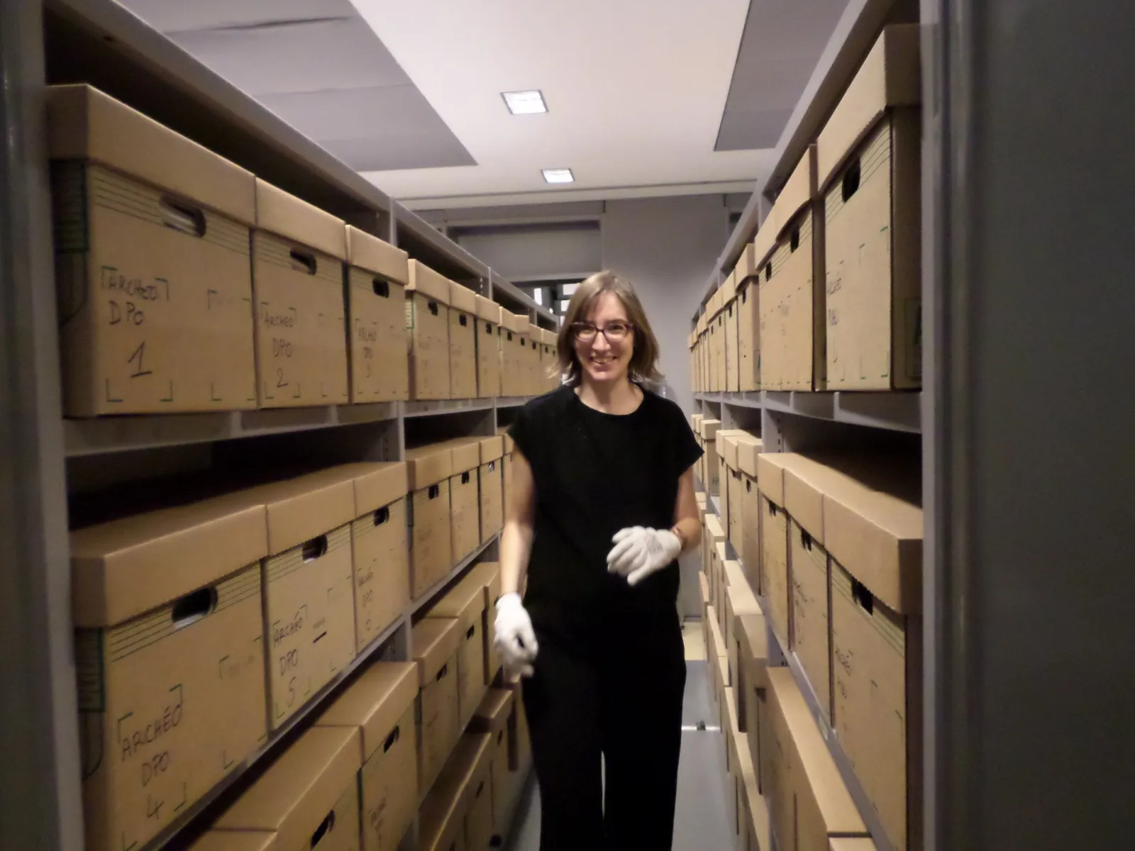 Vanessa Noizet gantée dans les réserves d’archives, entre les rayonnages de boîtes.